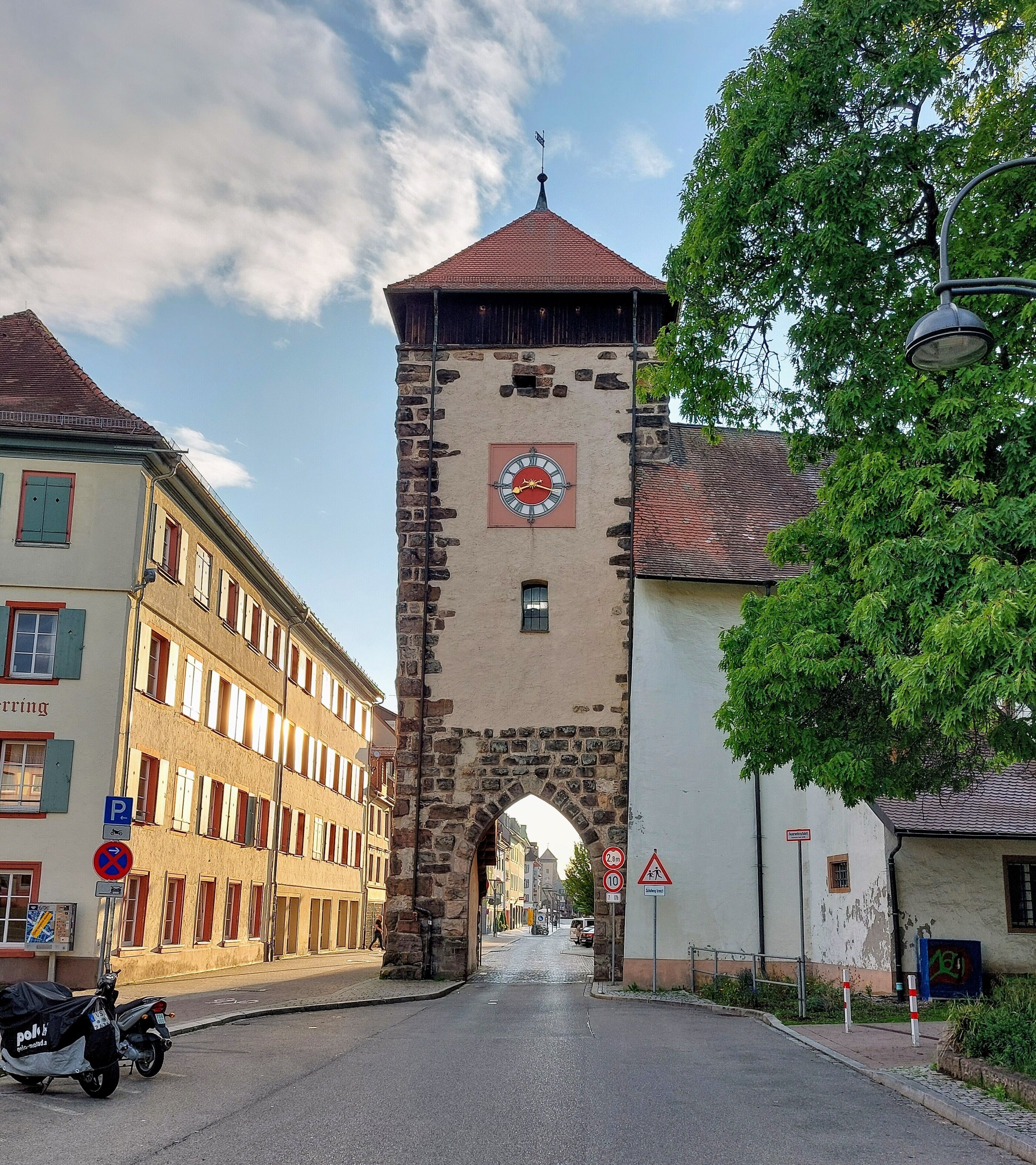 A street with a historic tower featuring a clock, a clear sky, and a tree.
