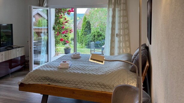 A bedroom with a bed, a chair, a bedside table, a TV, and a view of a garden through a sliding door.
