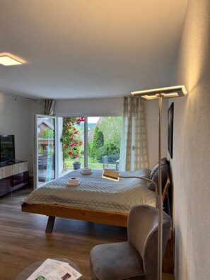 Un dormitorio con cama, una silla, una mesita de noche, televisión y vista a un jardín por una puerta corredera.