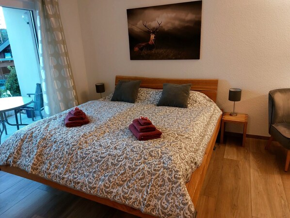 Un dormitorio con edredón estampado, dos almohadas, cabecera de madera, mesita de noche con lámpara y una silla.