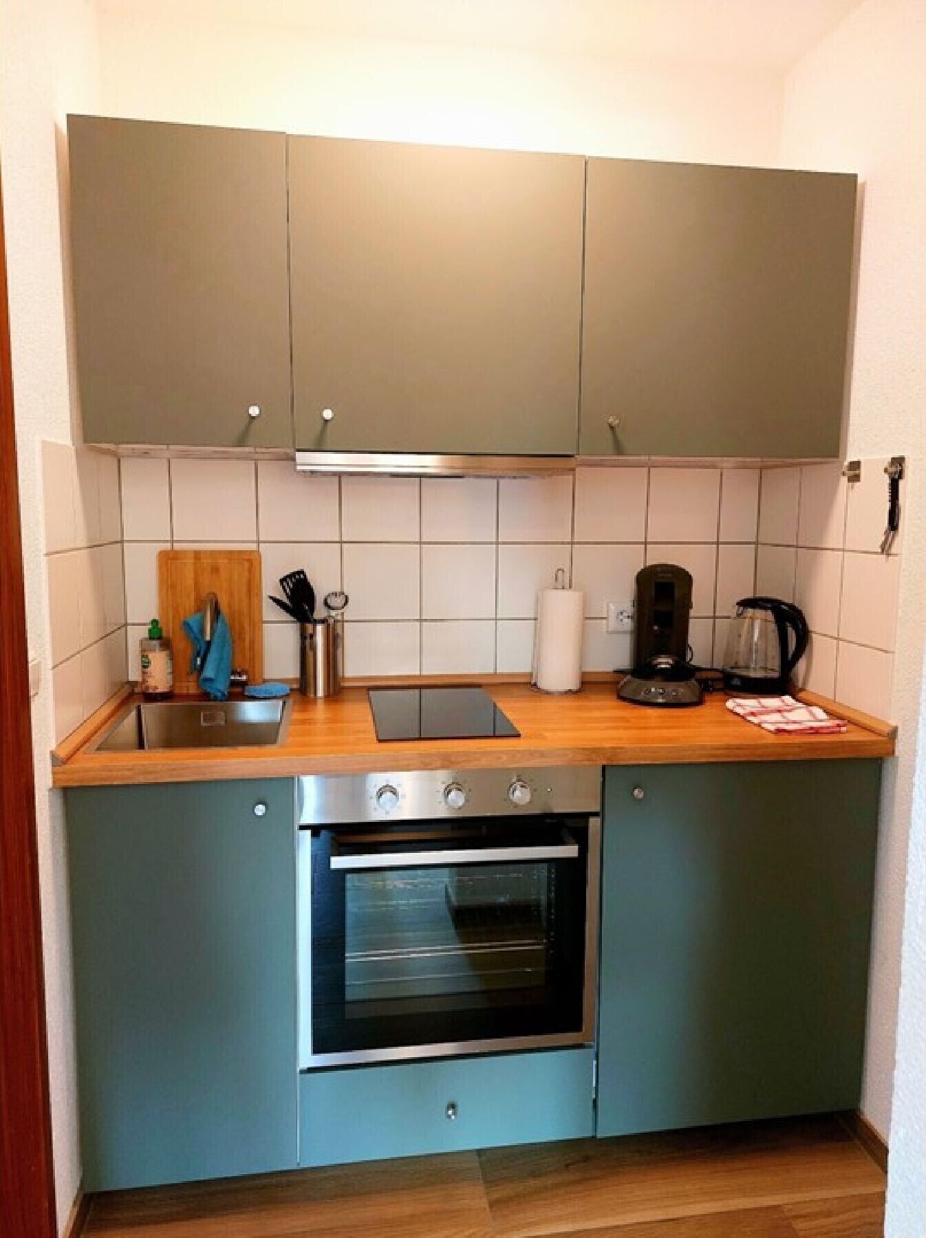 Eine kleine Küche mit grauen Schränken, einem Edelstahlbackofen, einer Kaffeemaschine und einer Holz Arbeitsplatte.