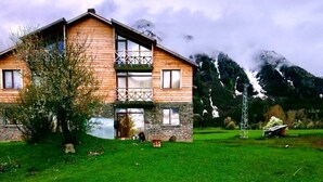 A two-story wooden house with a balcony, surrounded by a green lawn and mountains in the background.