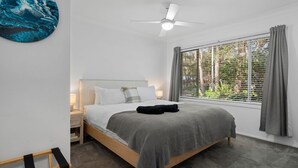 Ein Schlafzimmer mit Bett, Nachttisch, Deckenventilator, Fenster mit Vorhängen und Blick auf Grünfläche.