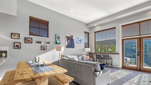 A modern living room with a wooden table, grey sofa, and large windows with blinds.