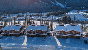 A snow-covered residential area with multiple houses, each with a distinct architectural style, surrounded by a forested mountain backdrop. - Modern Open Concept 2 bedroom Ski In/Ski Out Bottom Floor Unit! (Sun Peaks)