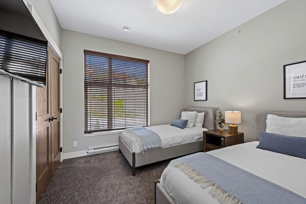 A bedroom with two beds, a window with blinds, a bedside table with a lamp, and a framed quote on the wall.