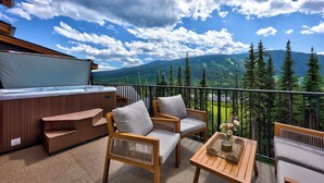 A balcony with wooden furniture, a hot tub, and a view of mountains and trees.