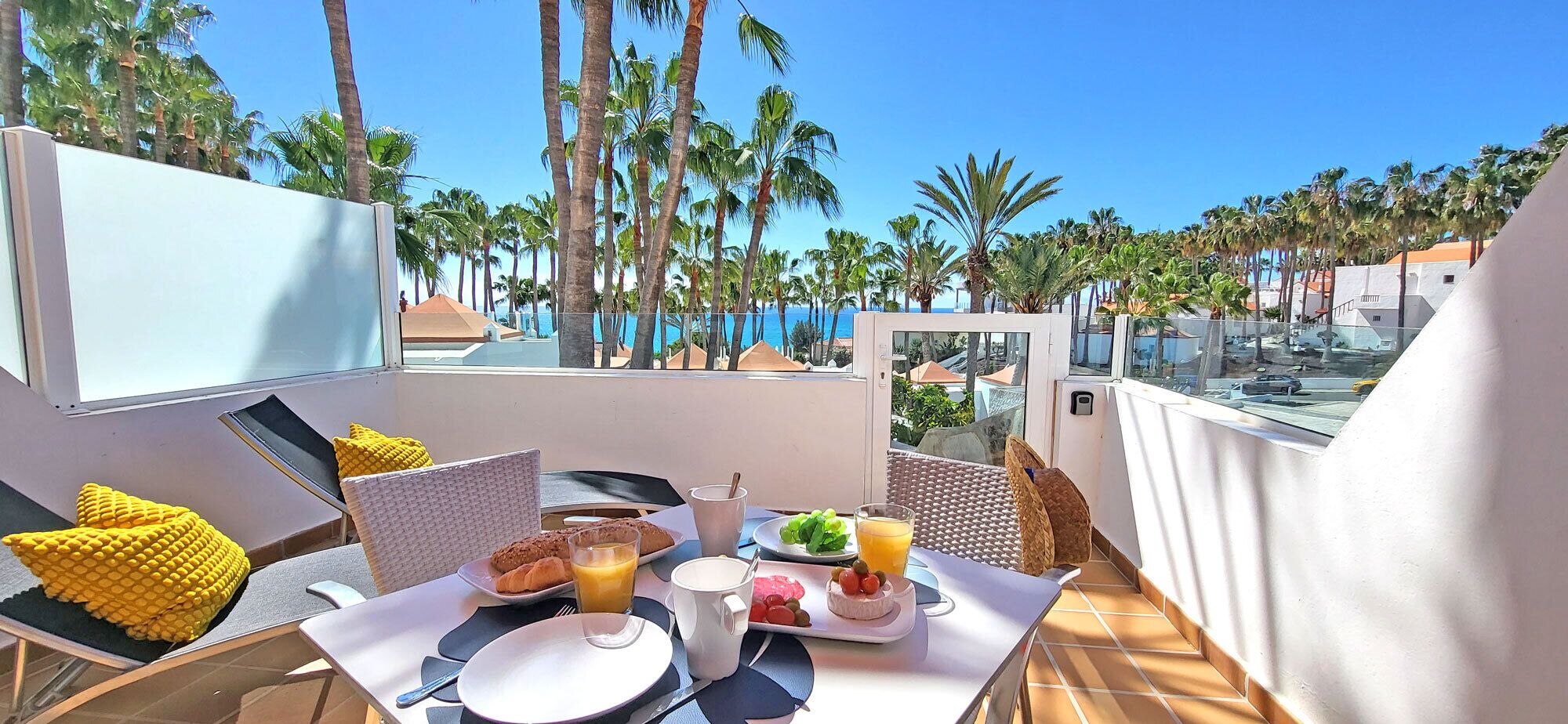 A balcony with a table set for breakfast, overlooking a pool and palm trees.