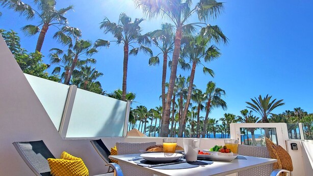 A sunlit outdoor dining area with a table set for two, overlooking a beach with palm trees and the ocean. - Vacation apartment EL PARAISO in Costa Calma, close to the beach, pool, terrace (Costa Calma)