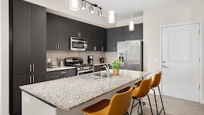 A modern kitchen with dark cabinets, stainless steel appliances, a granite countertop, and yellow bar stools.