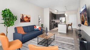 A modern living room with a blue sofa, orange armchairs, and a coffee table. A kitchen is visible in the background with dark cabinets and a white island.