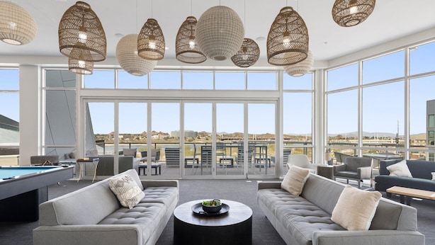 A spacious lounge area with a pool table, grey sofas, and a round coffee table. Large windows offer a view of the cityscape.