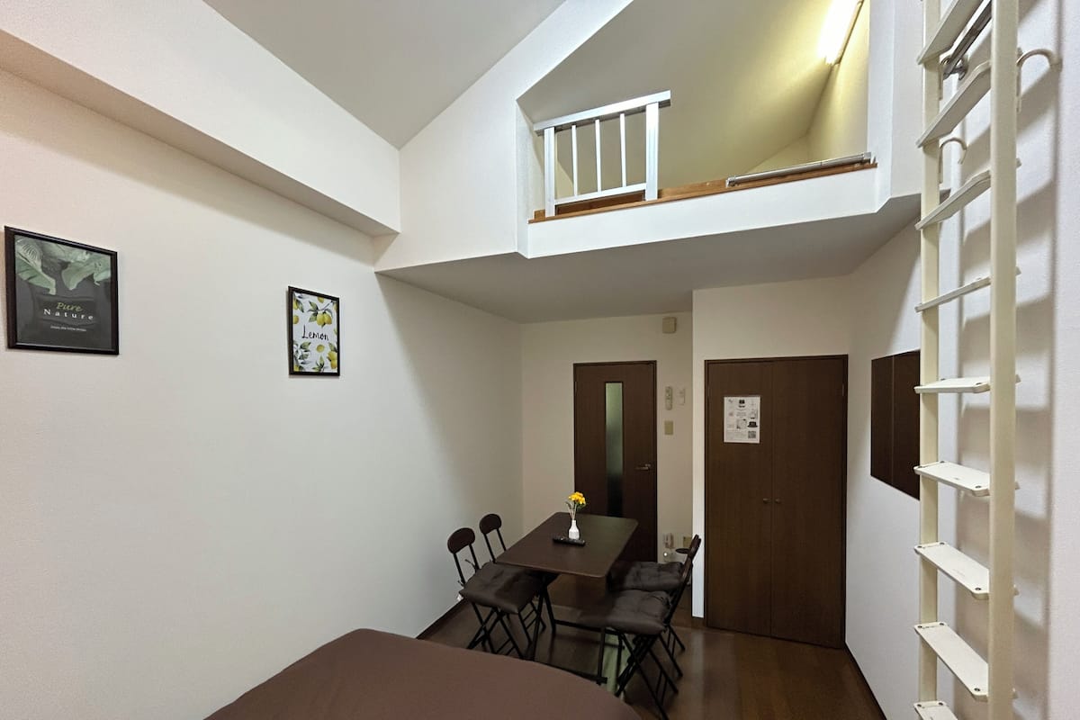 A compact room with a loft bed, a dining table with chairs, and a small balcony.