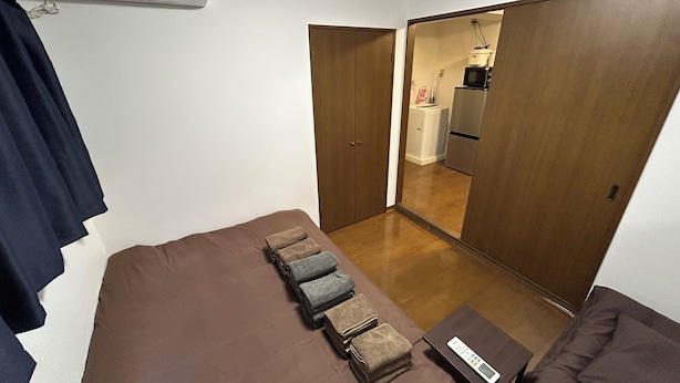 A small, compact room with a bed, a bedside table with a remote control, and a set of towels. There is a wardrobe and a door leading to a kitchenette.
