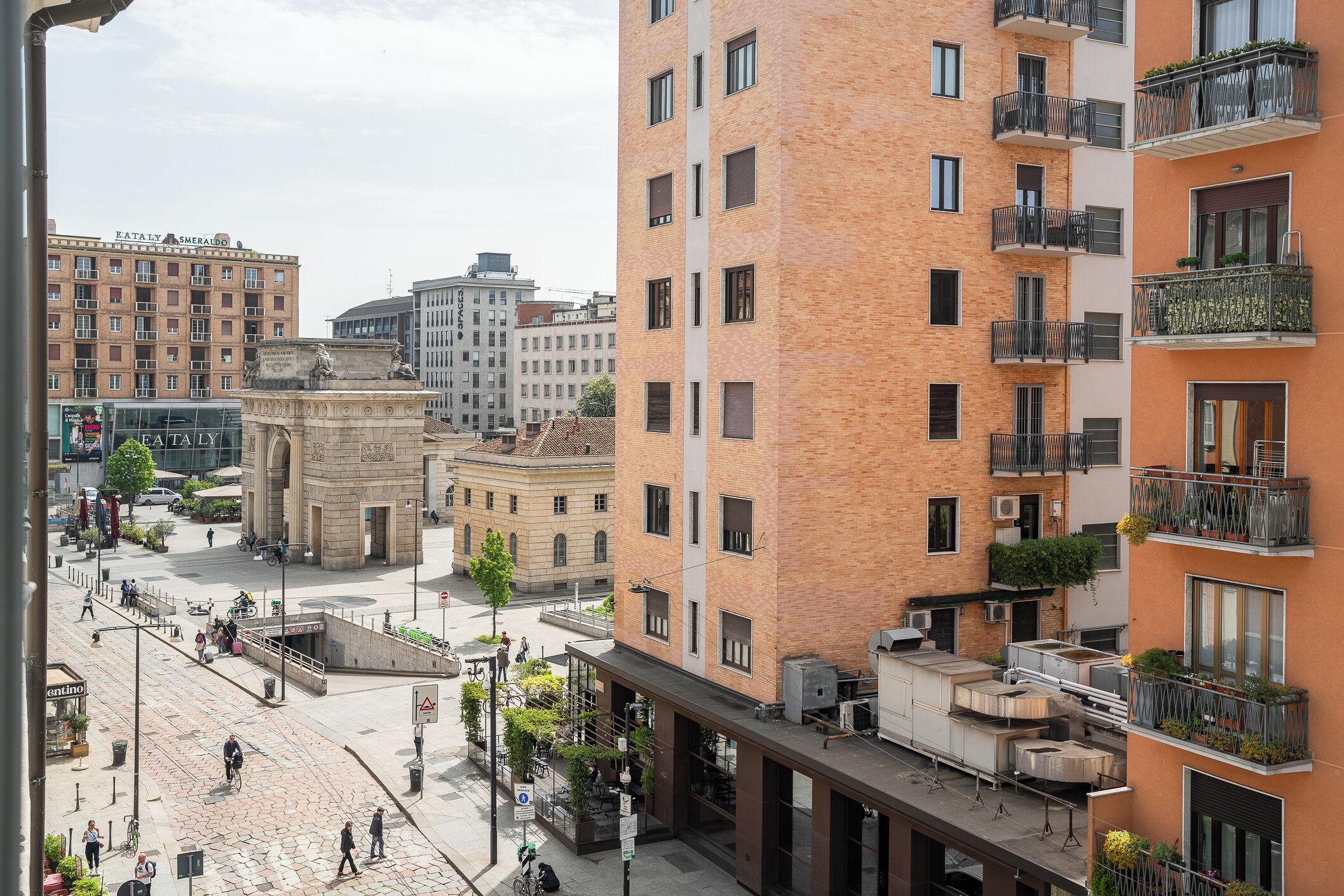 Un immeuble à plusieurs étages doté de balcons, une place centrale ouverte et des bâtiments urbains environnants.