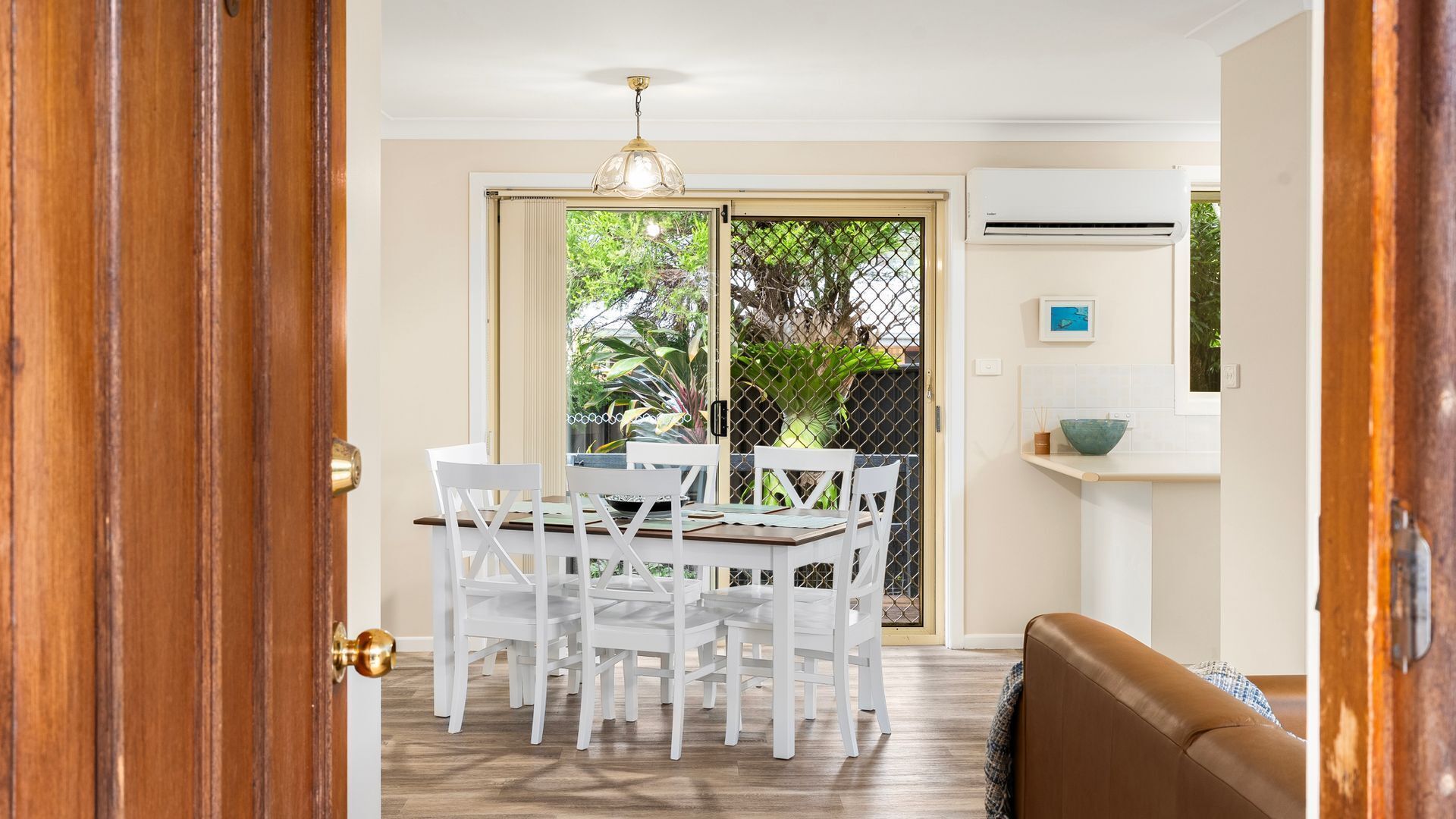 A dining area with a table and chairs, a ceiling light, a sliding glass door leading to a garden, and a brown couch in the foreground.
