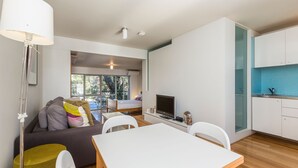 A modern living room with a sofa, a dining table, and a TV. There is a kitchen area with a blue backsplash and white cabinets. - Cottesloe Tree Top Studio (Cottesloe)