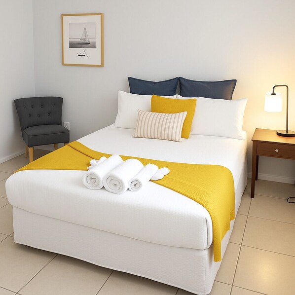 Uma cama bem arrumada com roupa de cama branca e amarela, uma poltrona cinza, uma mesa de cabeceira com abajur e um quadro emoldurado na parede.