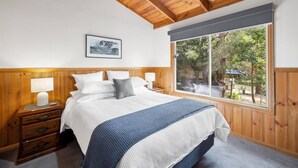 Een slaapkamer met een houten hoofdbord, een groot raam met uitzicht op groen, een nachtkastje met een lamp en een lijst met een foto aan de muur.