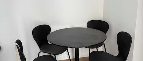 Una mesa redonda con cuatro sillas negras en una esquina de una habitación con piso de madera y paredes blancas.