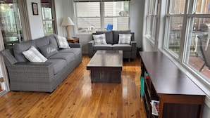 Un salon avec deux canapés gris, une table basse en bois et une grande fenêtre donnant sur une maison et des arbres.