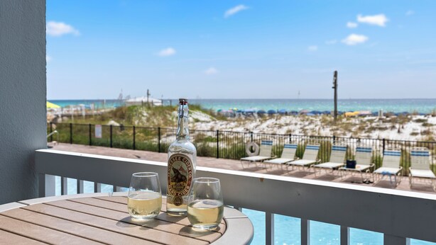 A balcony with a view of the beach and ocean. There is a bottle of liquor and two glasses on the table.