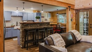 A cozy cabin interior with a stone fireplace, wooden ceiling, and a dining area with a red chair.