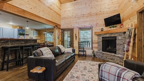 A cozy cabin interior with a stone fireplace, a black leather sofa, a wooden dining table, and a kitchen area with a bar.