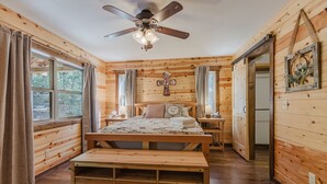 A wooden cabin bedroom with a bed, bedside tables, a bench, a ceiling fan, and a cross on the wall.