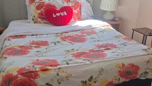 Una cama bien tendida con una colcha estampada con motivos florales y una almohada con forma de corazón rojo que dice 'LOVE'.