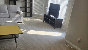 Una sala de estar con un sofá gris, una mesa de centro amarilla, un televisor sobre un mueble bajo y una estantería.