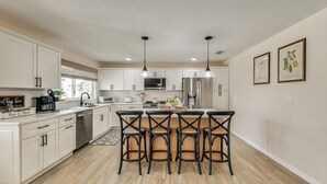 A modern kitchen with white cabinets, stainless steel appliances, a central island with bar stools, pendant lighting, and framed botanical artwork on the wall. - Private Pool w/ Canal Views: Apollo Beach Getaway (Apollo Beach)