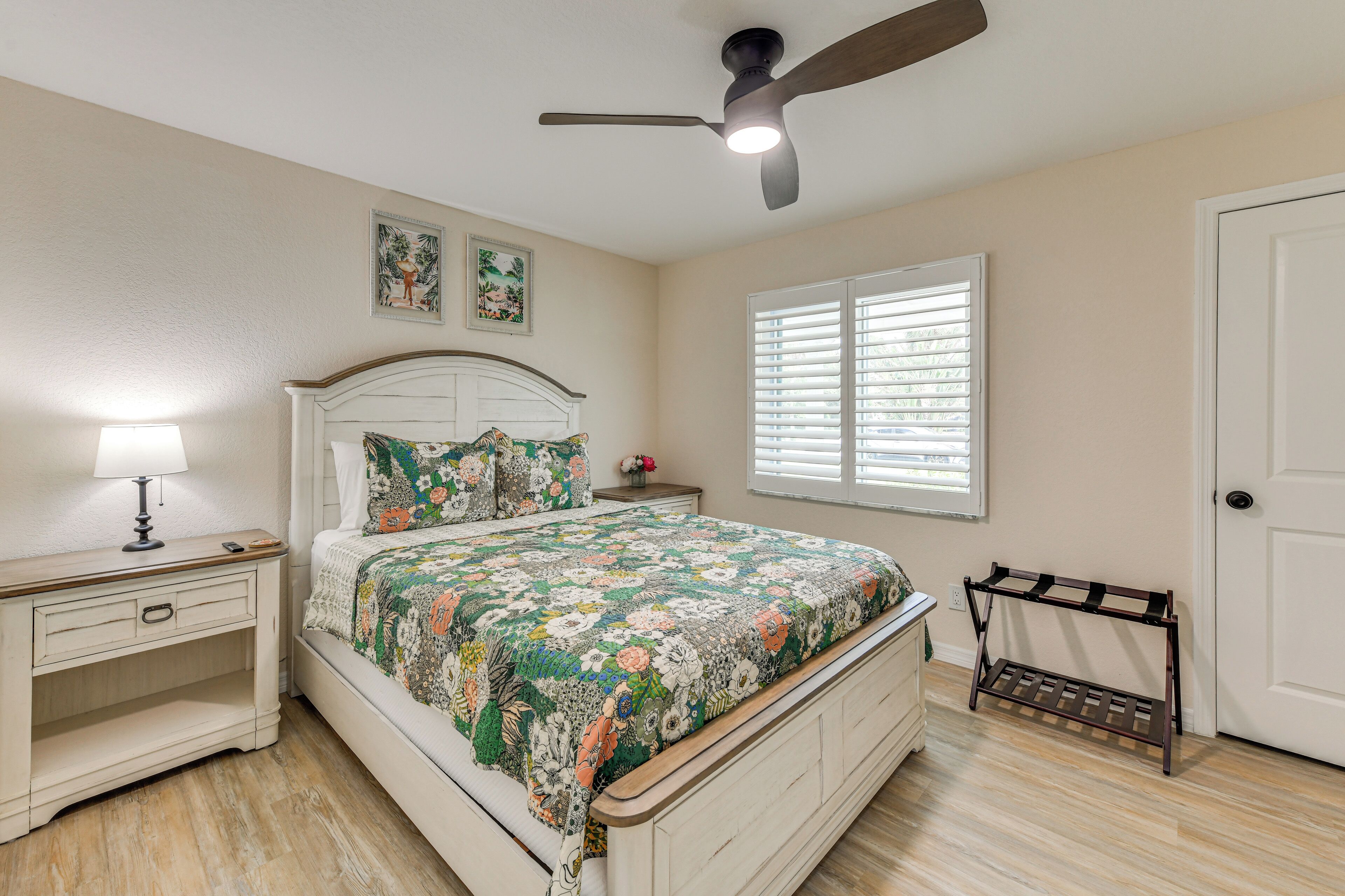 A bedroom with a bed, nightstand, lamp, ceiling fan, window with blinds, and a door.