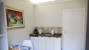 Une cuisine compacte avec un réfrigérateur blanc, un évier et un mur recouvert de carreaux. On y trouve une table à manger avec une nappe bleue et blanche, ainsi qu’une chaise. Un tableau est accroché au mur, au-dessus du comptoir.