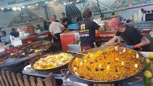 Un stand de nourriture avec de grandes poêles remplies de paella et de viandes grillées, un homme qui sert les plats, et une fresque colorée en arrière-plan.
