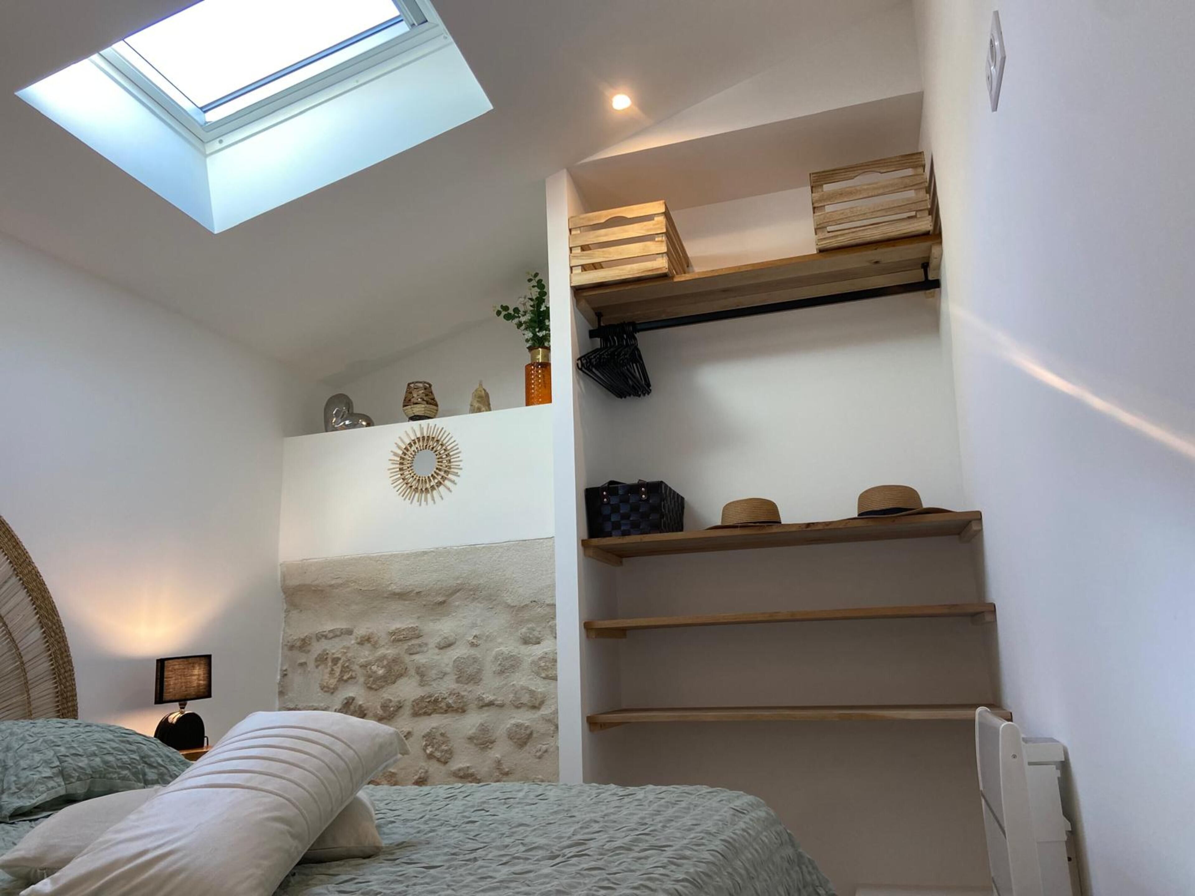 A bedroom with a skylight, a bed with pillows, a nightstand, a wall-mounted shelf with decorative items, and a radiator.