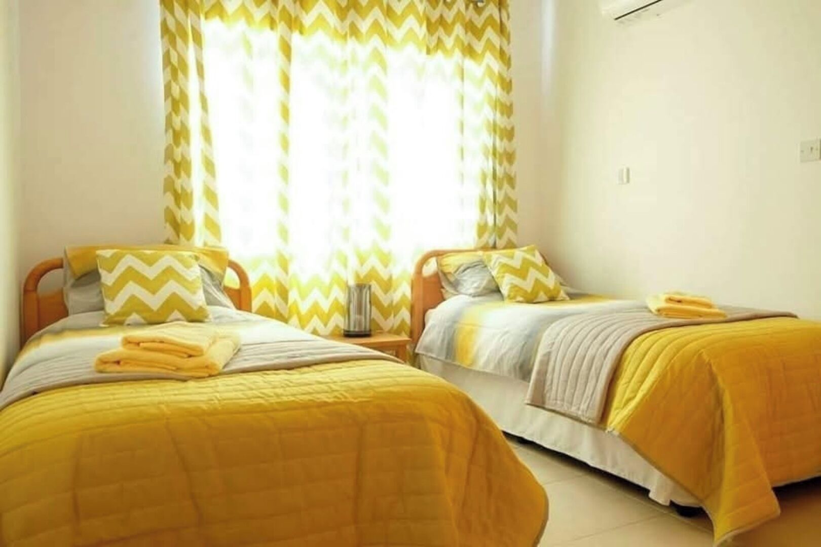 Una habitación con dos camas, cada una con ropa de cama amarilla y almohadas a juego. Hay una ventana con cortinas amarillas y blancas, y una mesita con una lámpara entre las camas.