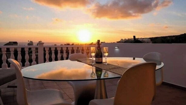 A round glass table with four chairs on a terrace, set for a meal with a bottle and glasses, overlooking a sunset and cityscape.