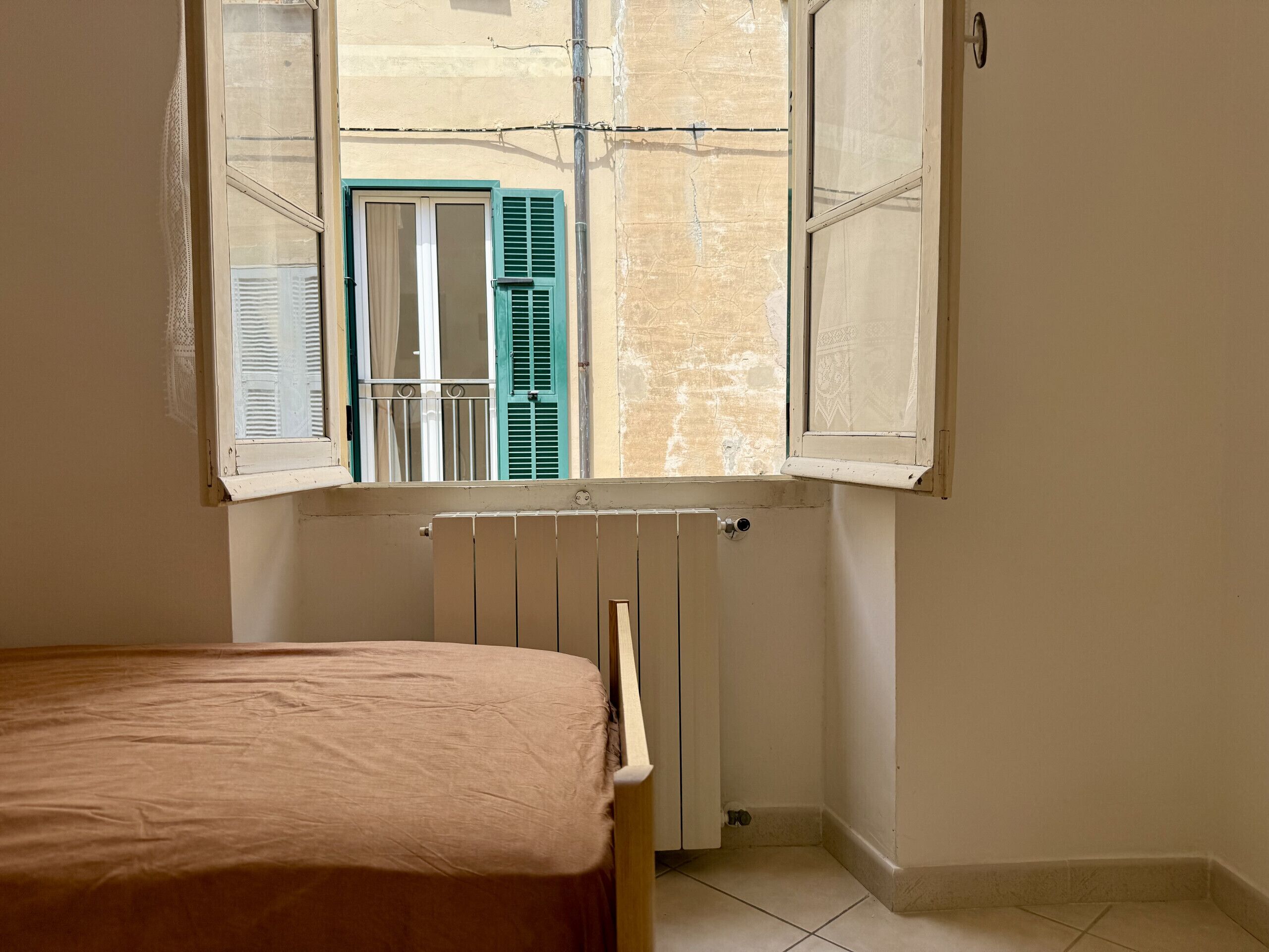Une chambre avec une fenêtre donnant sur la rue, un radiateur et un lit recouvert d’une couverture beige.