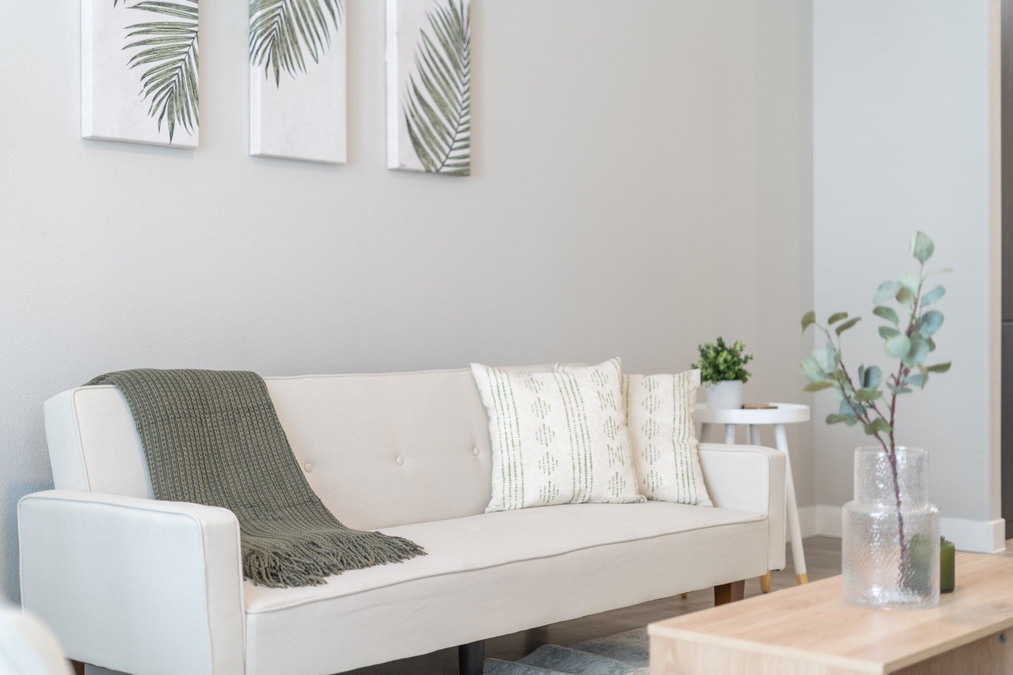 A modern living room with a white sofa, green throw blanket, and decorative pillows. There is a small white table with a plant and a vase.