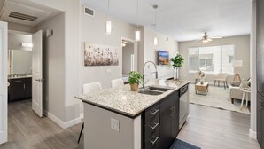 A modern kitchen with a granite countertop, dark cabinetry, and a central island. There is a dining area with a white table and chairs, and a large window with blinds. - Luxury Tempe Stay 1BR w/ Patio, Gym & Business Center (Tempe)
