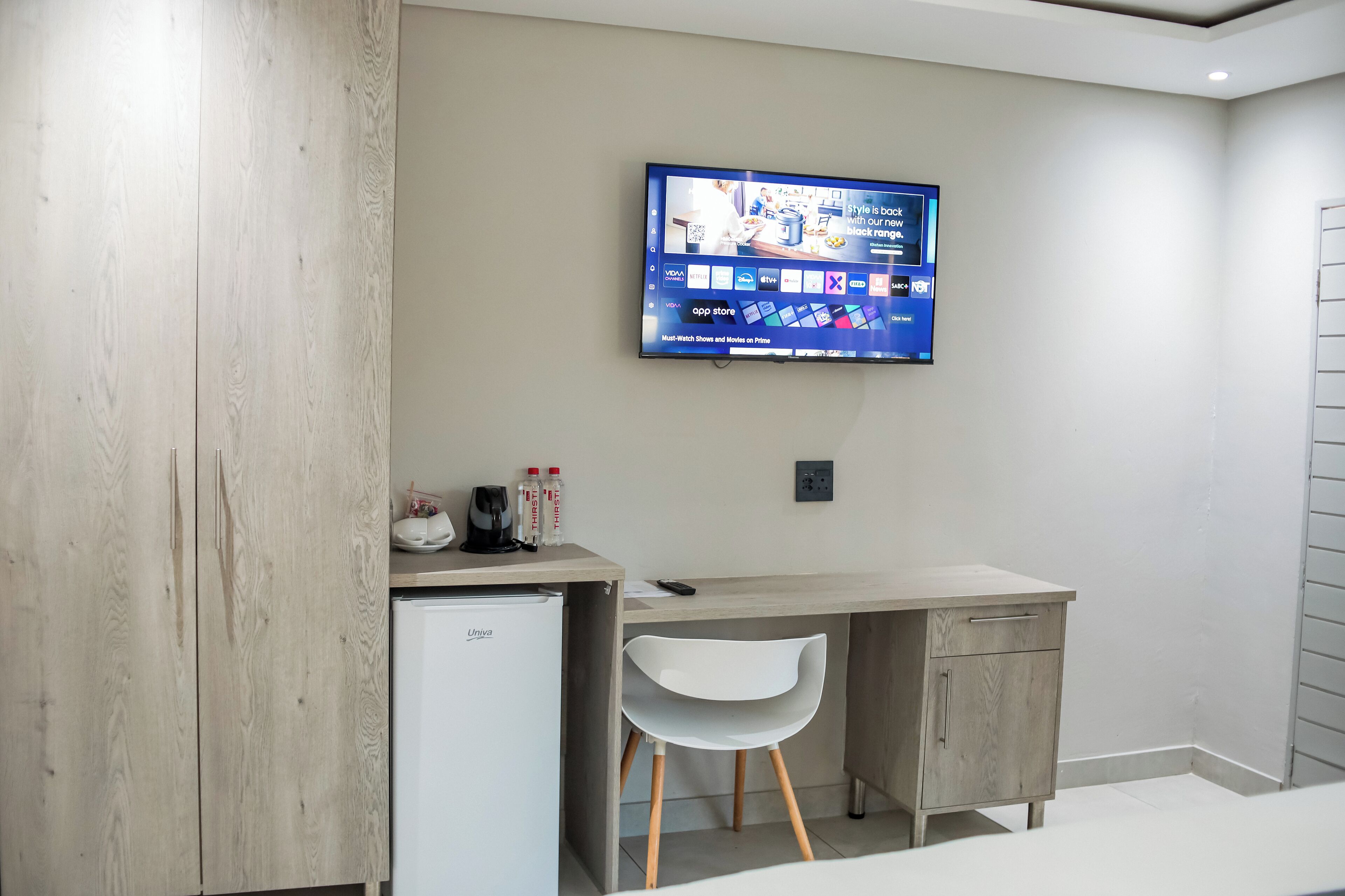 Quarto moderno com televisão de tela plana na parede, uma escrivaninha de madeira com cadeira branca e um refrigerador pequeno.