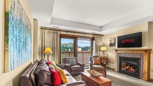 Ein Wohnzimmer mit einer Couch, Sesseln, einem Kamin, einem Fernseher und einem großen Fenster mit Blick auf die Berge.