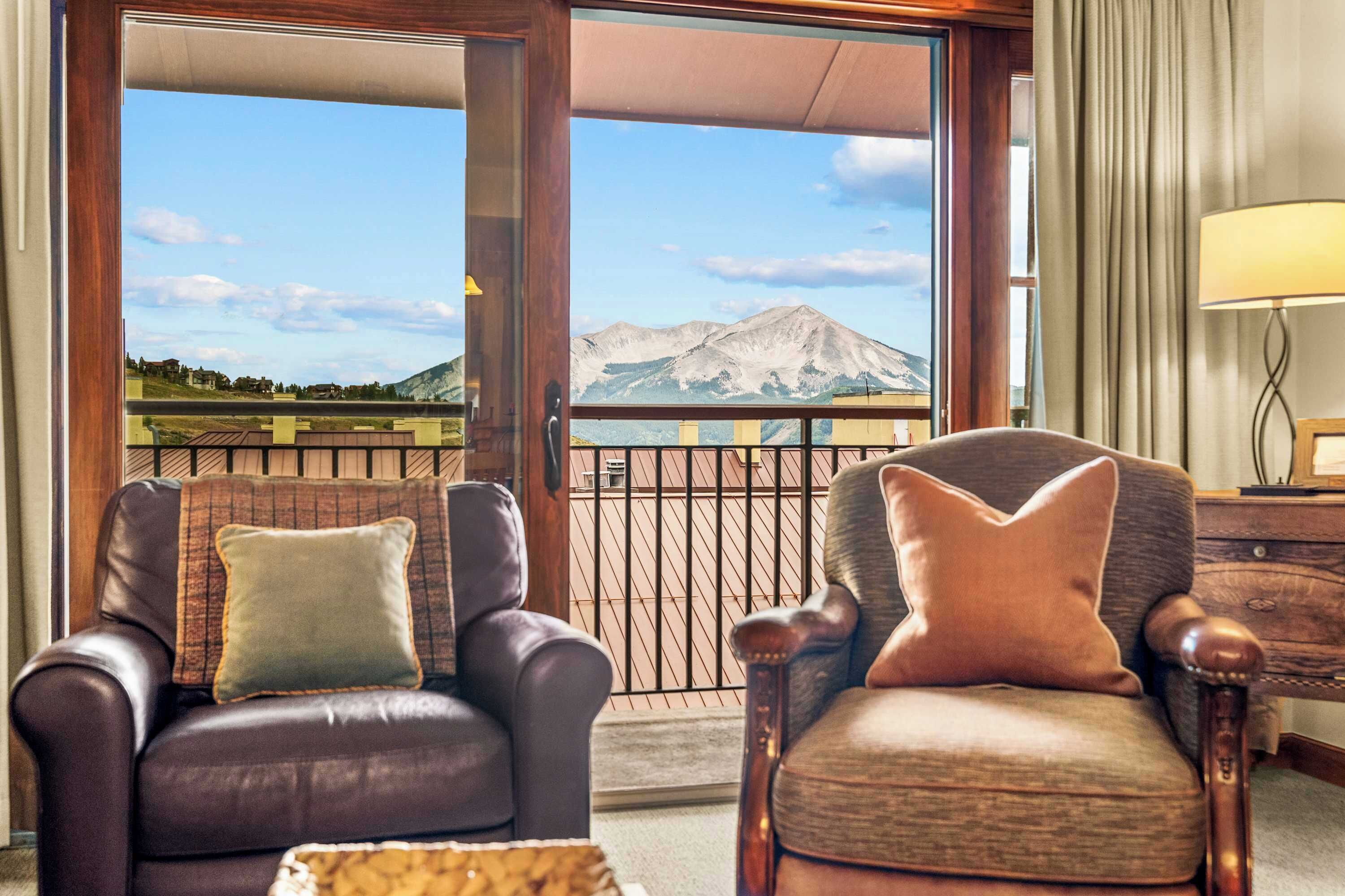 A room with two leather armchairs, a wooden side table, and a view of mountains through large windows.