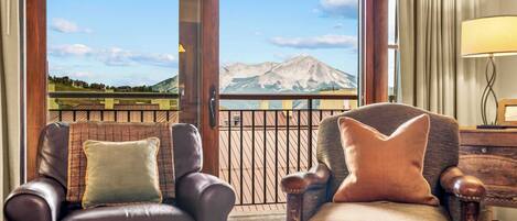 A room with two leather armchairs, a wooden side table, and a view of mountains through large windows.
