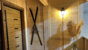 Un intérieur en bois avec une porte, des skis appuyés contre le mur, une lampe et un vase contenant des plantes séchées.