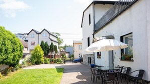 A courtyard with a paved walkway, outdoor seating area with tables and chairs, and a white building with a balcony. - La Perla (Międzyzdroje)