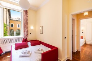 A bedroom with a red bed, white linens, a window with a view of buildings, and a wooden floor. - Rental in Rome Baccina Apartment (Rome)
