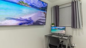 Quarto com uma televisão grande fixada na parede exibindo uma cena de praia, um monitor menor em uma mesa, uma cadeira de escritório preta e uma janela com cortinas.