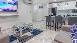 Uma sala de estar moderna com televisão, mesa de centro de vidro e sofá. Há uma área de cozinha com mesa e cadeiras.